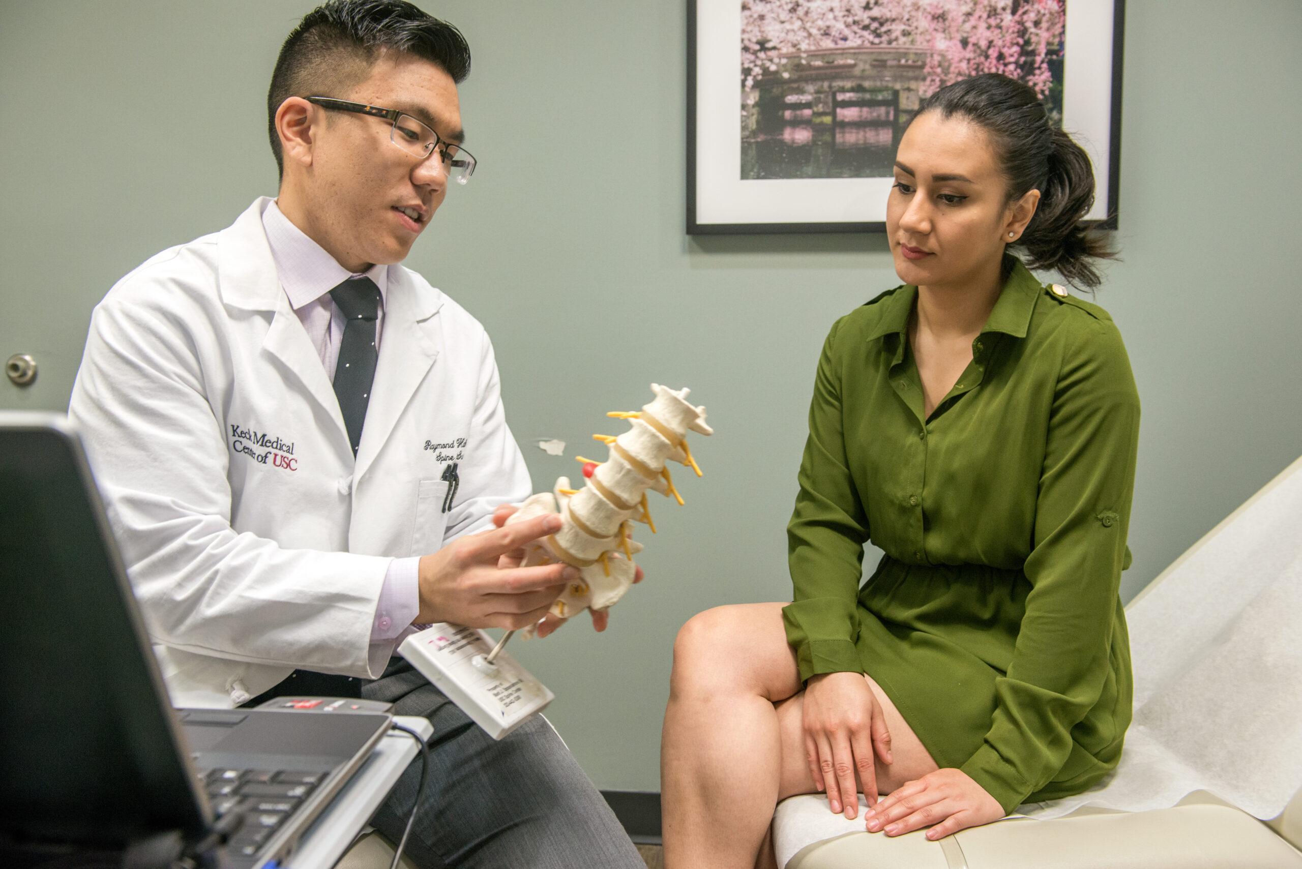 A male Keck Medicine laminectomy expert shows a female patient with back pain in a green dress a 3D model of a spine