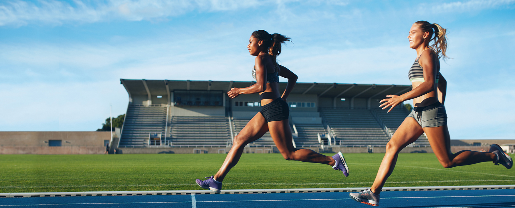 Two young female professional athletes in athletic wear and ponytails run on track in front of grass and bleachers