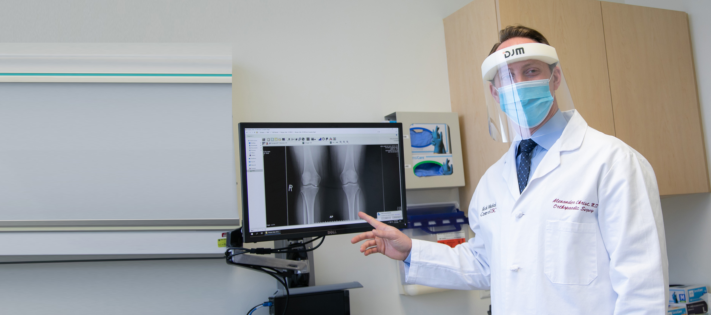 Dr. Alexander Christ, a Keck Medicine orthopedic surgeon in a mask and face shield, points to an X-ray of a knee replacement