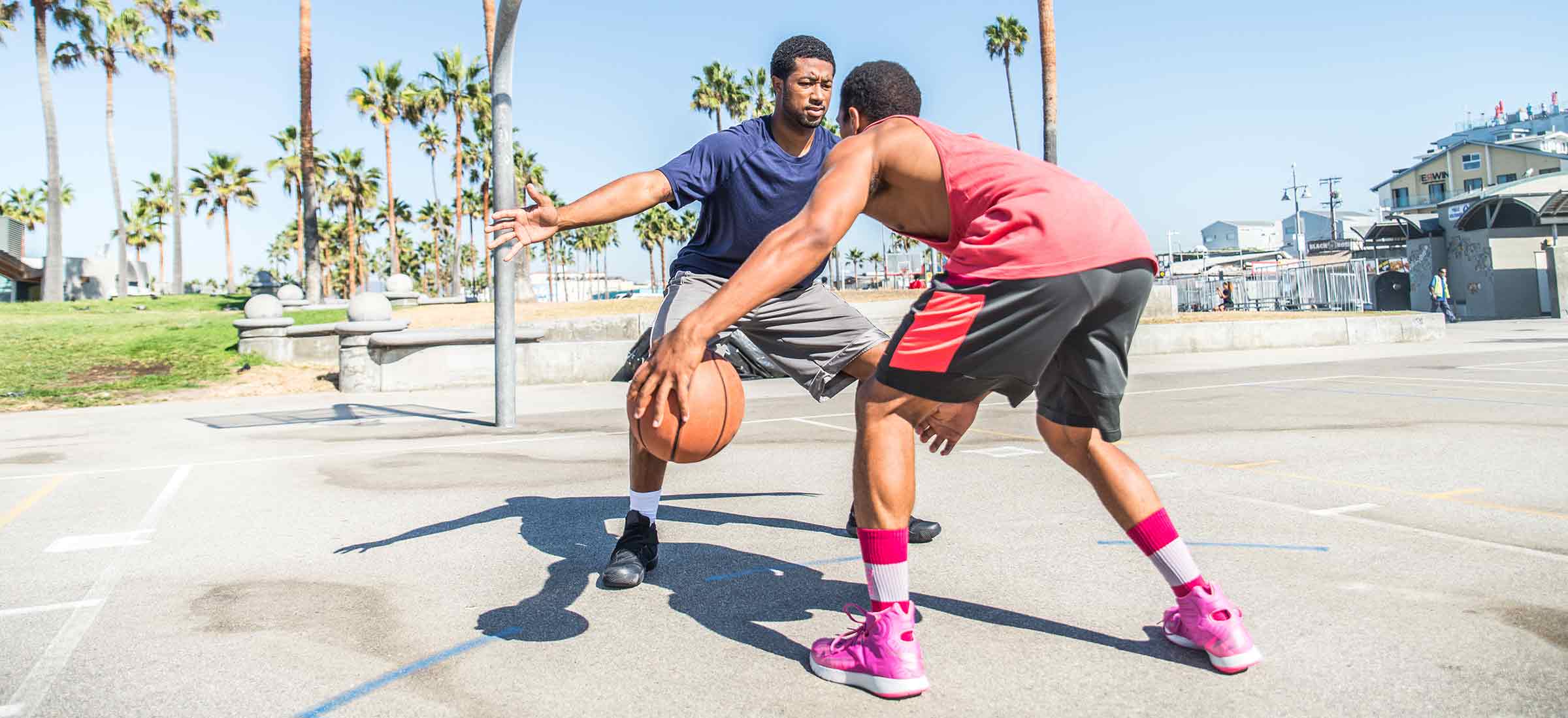 Two men play basketball on a court with palm trees in the background.