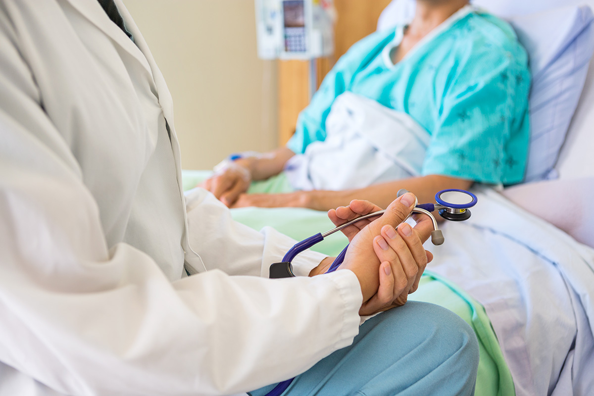 A doctor who checks for colon cancer sits at a patient's bedside, holding a stethoscope
