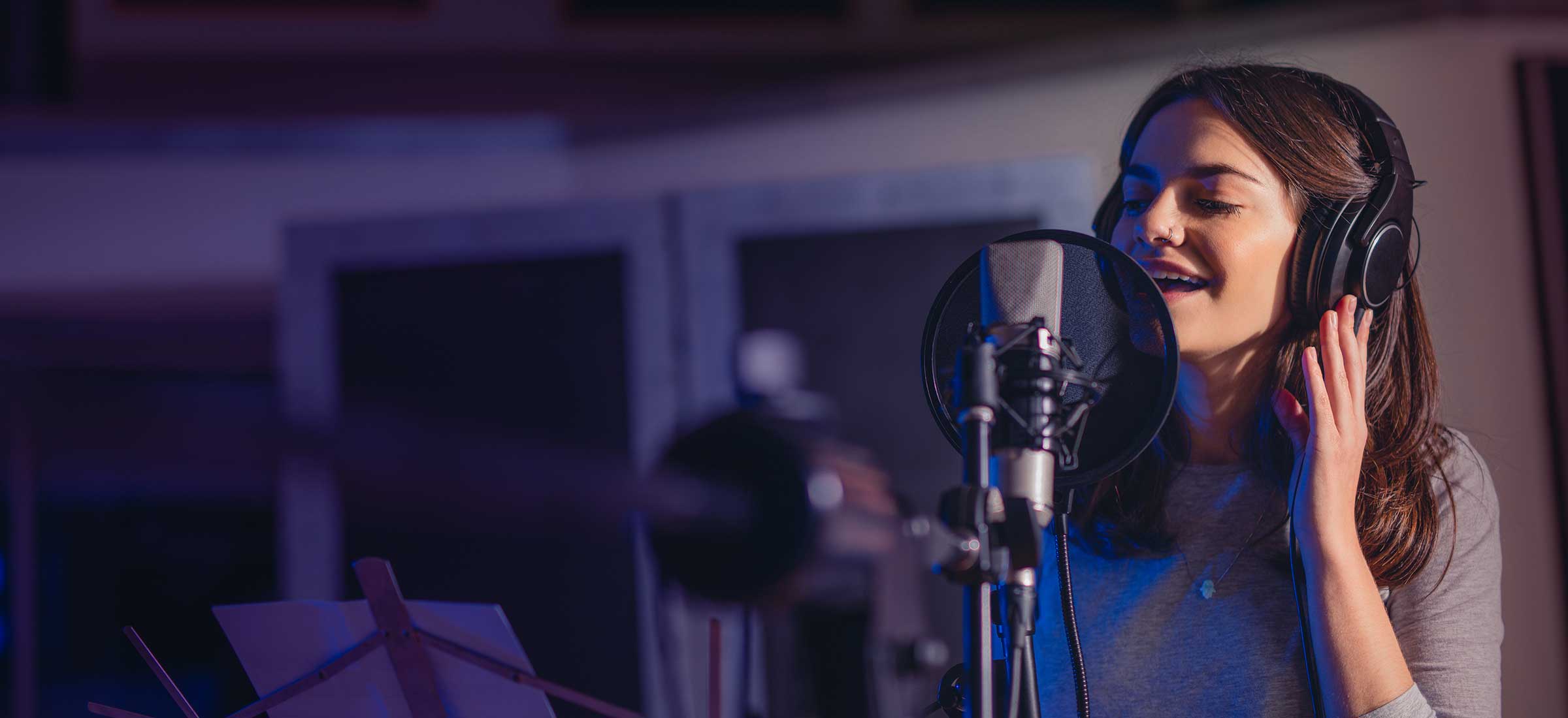 A woman sings into a microphone with headphones on in a studio.