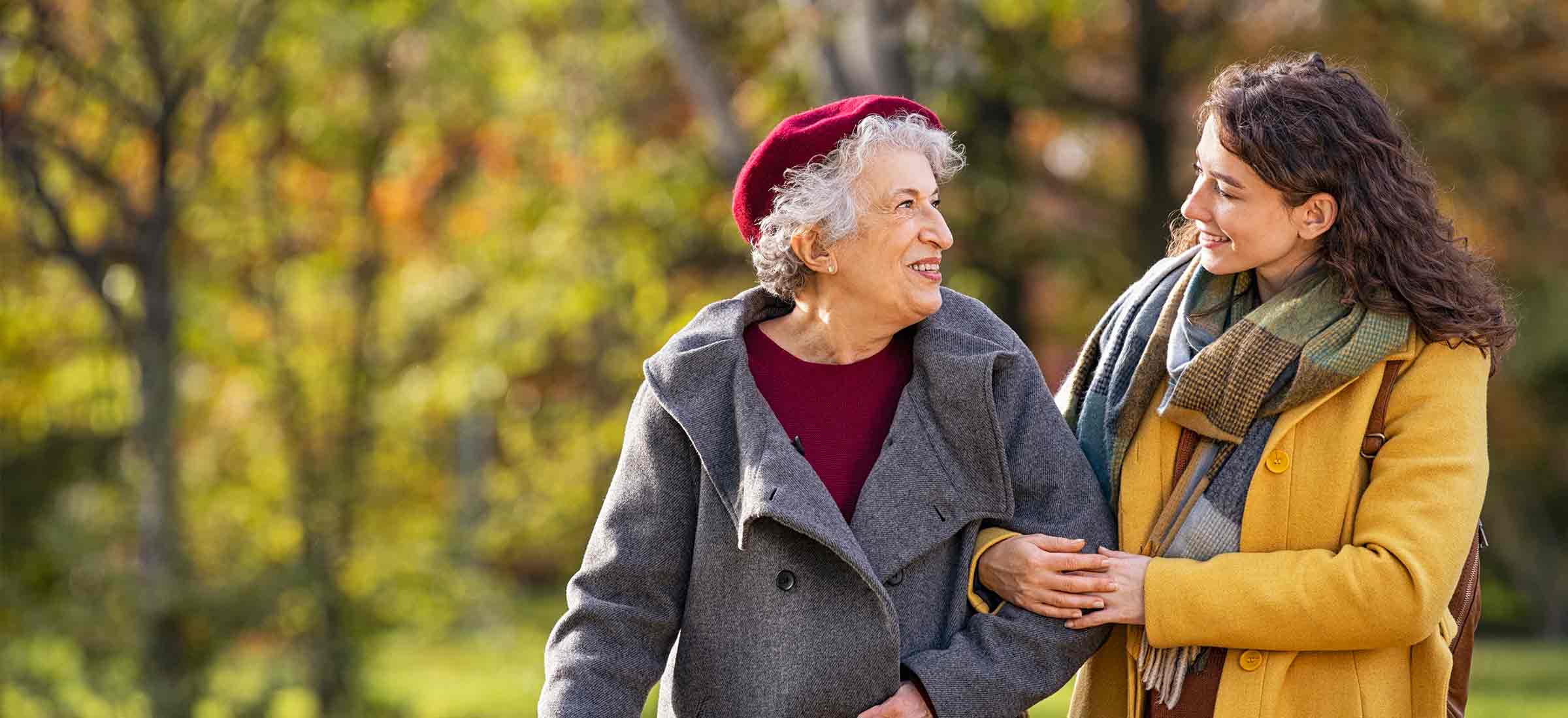 Two women smile at each other in coats while walking through a park.