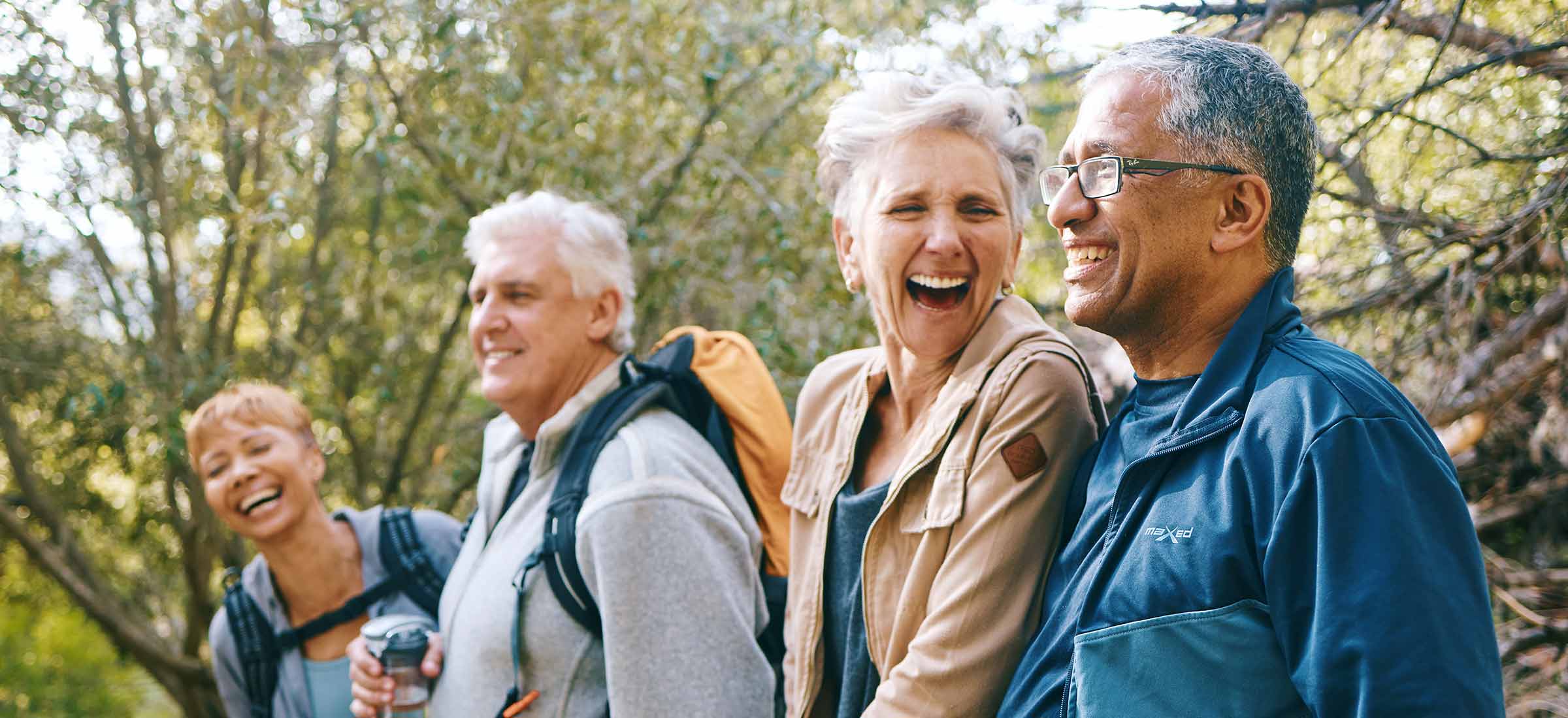 A group of elderly people laughs while walking through the woods wearing backpacks.
