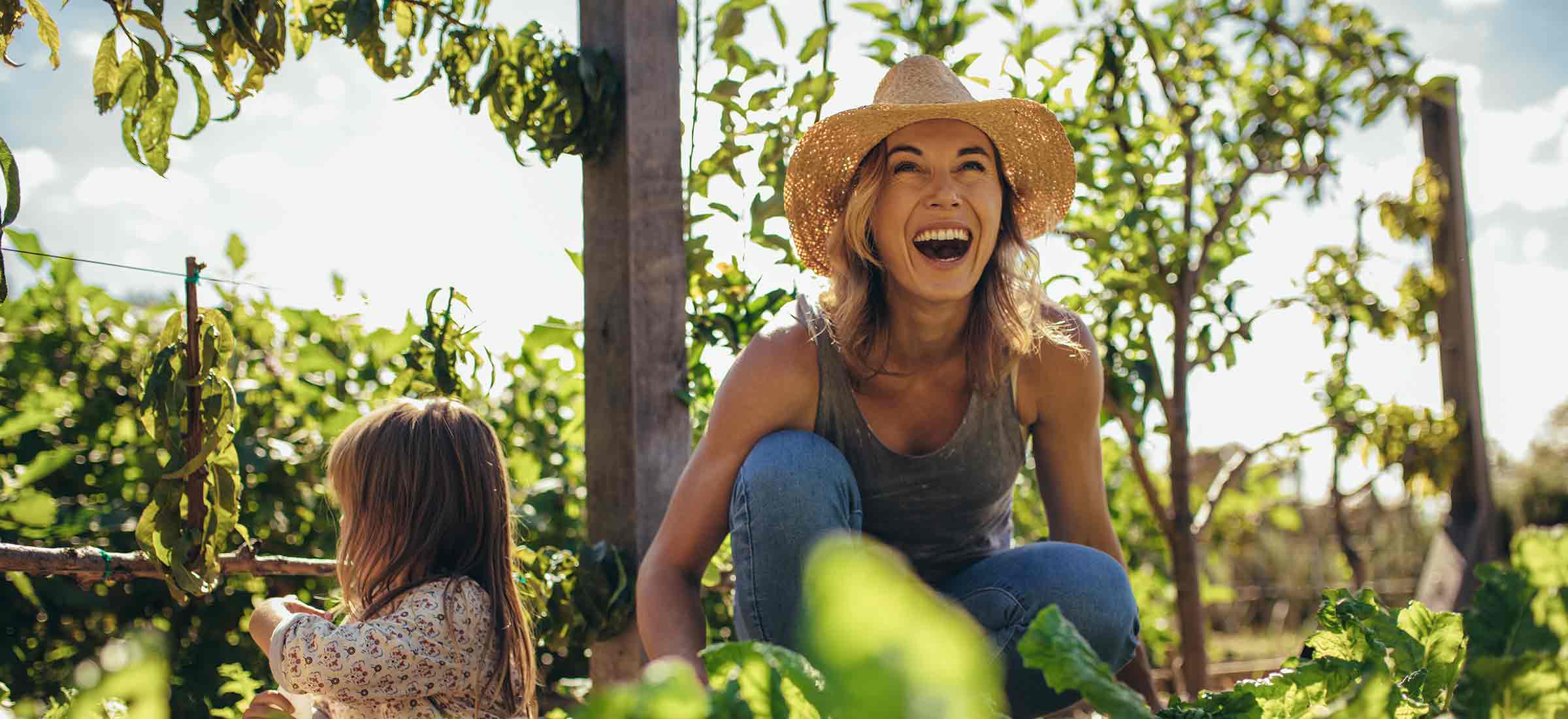 A woman wearing a hat smiles while she bends down in a garden next to a child.