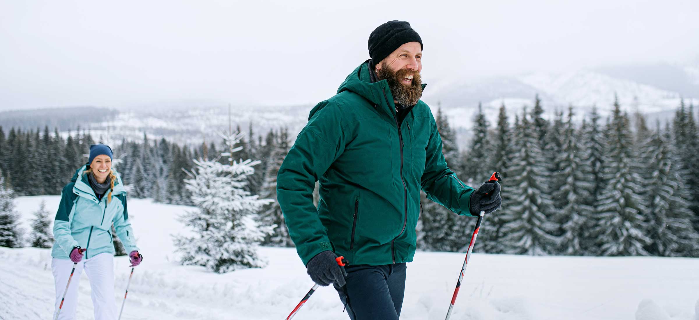 A man in a green coat with a beard cross country skis in a snowy forest with a woman.