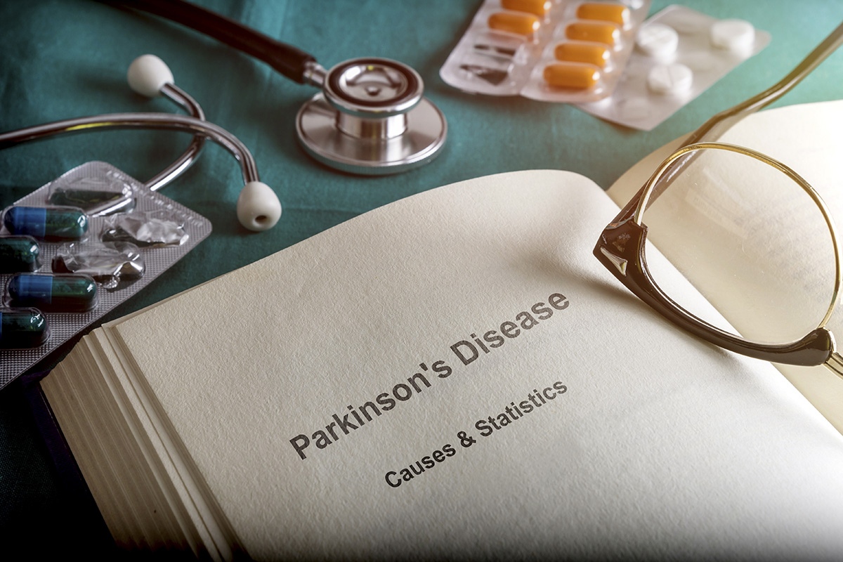 Eyeglasses sit atop an open book called Parkinson's Disease Causes & Statistics near a stethoscope, capsules and tablets
