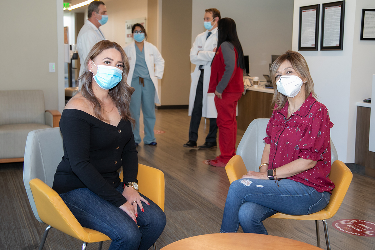 Two young women in masks sit together in yellow and white chairs in medical office, consulting with experts on liver disease
