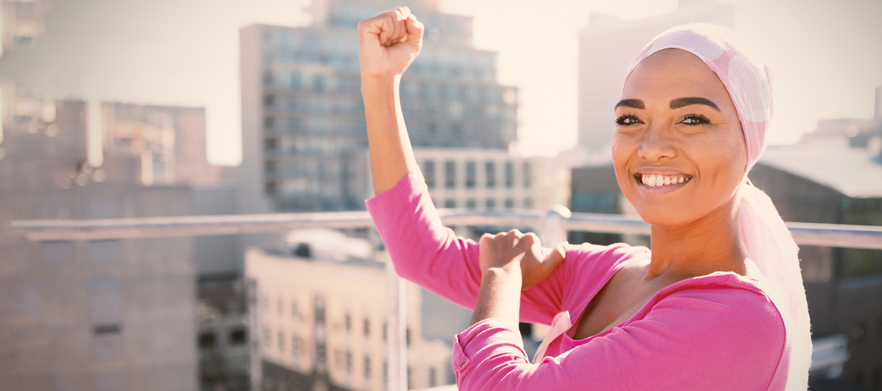 A young breast cancer patient wears a pink head scarf and shirt, holds her bent right arm, makes a winning fist and smiles