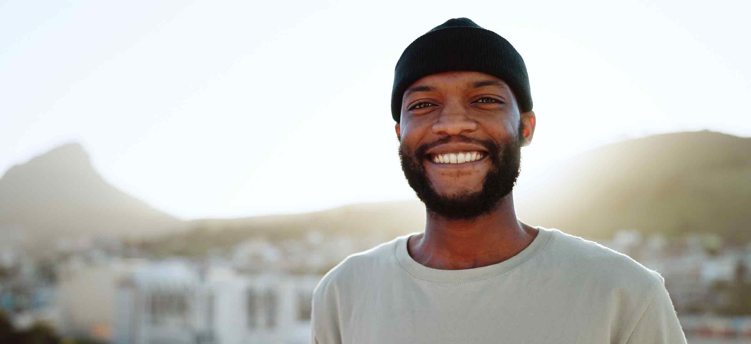 A black man smiles outdoors in a city with nature in the background during summer.