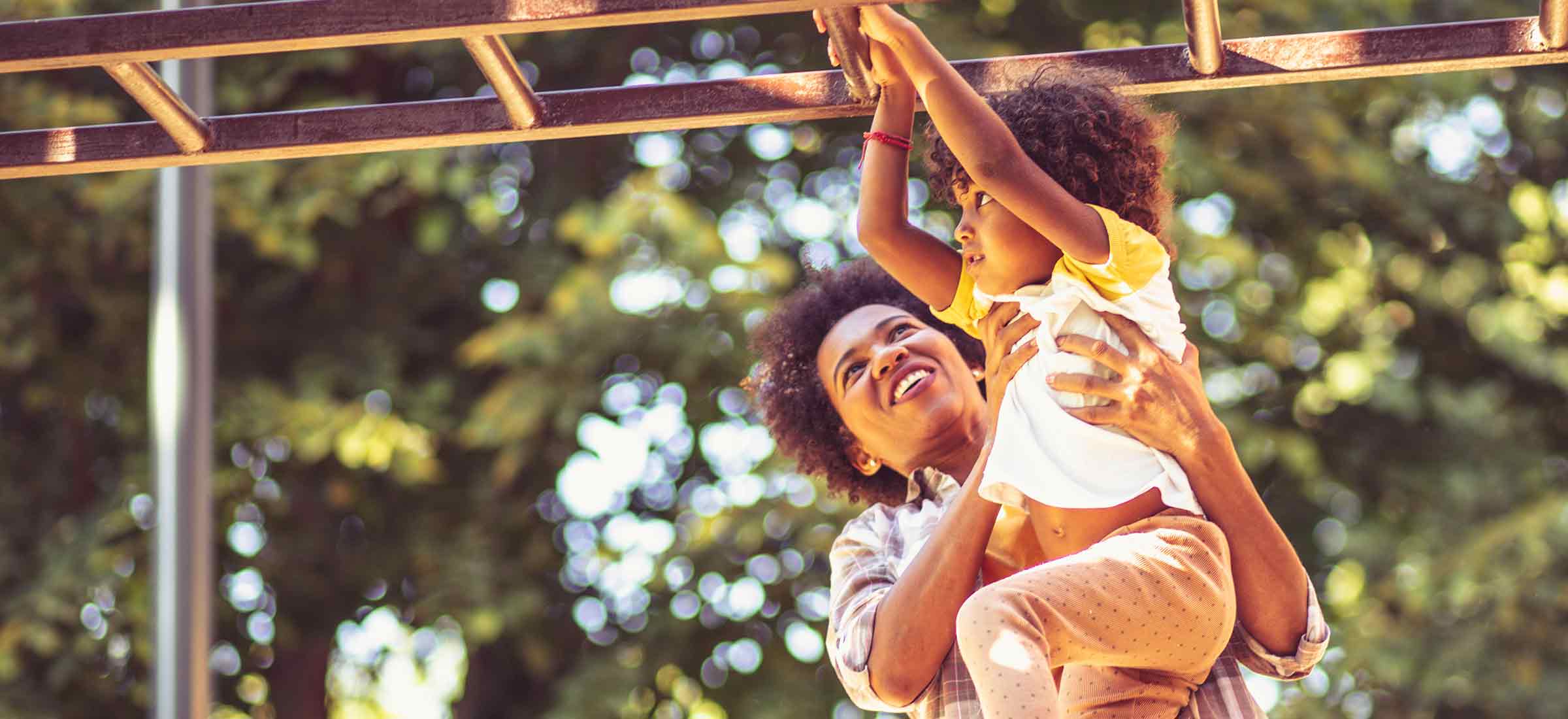 A woman holds a child up to assist them on the monkey bars on a playground.