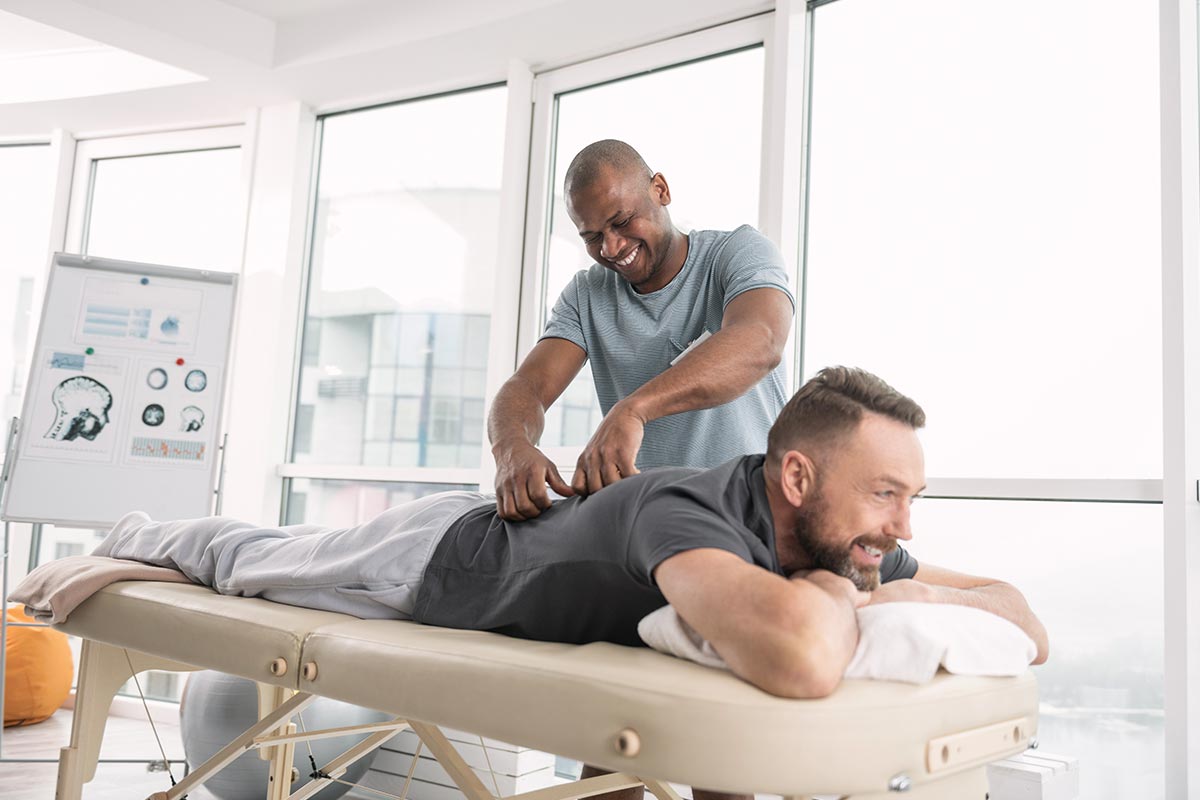 Happy delighted man smiling while giving a back massage for his patient in a relaxing atmosphere.