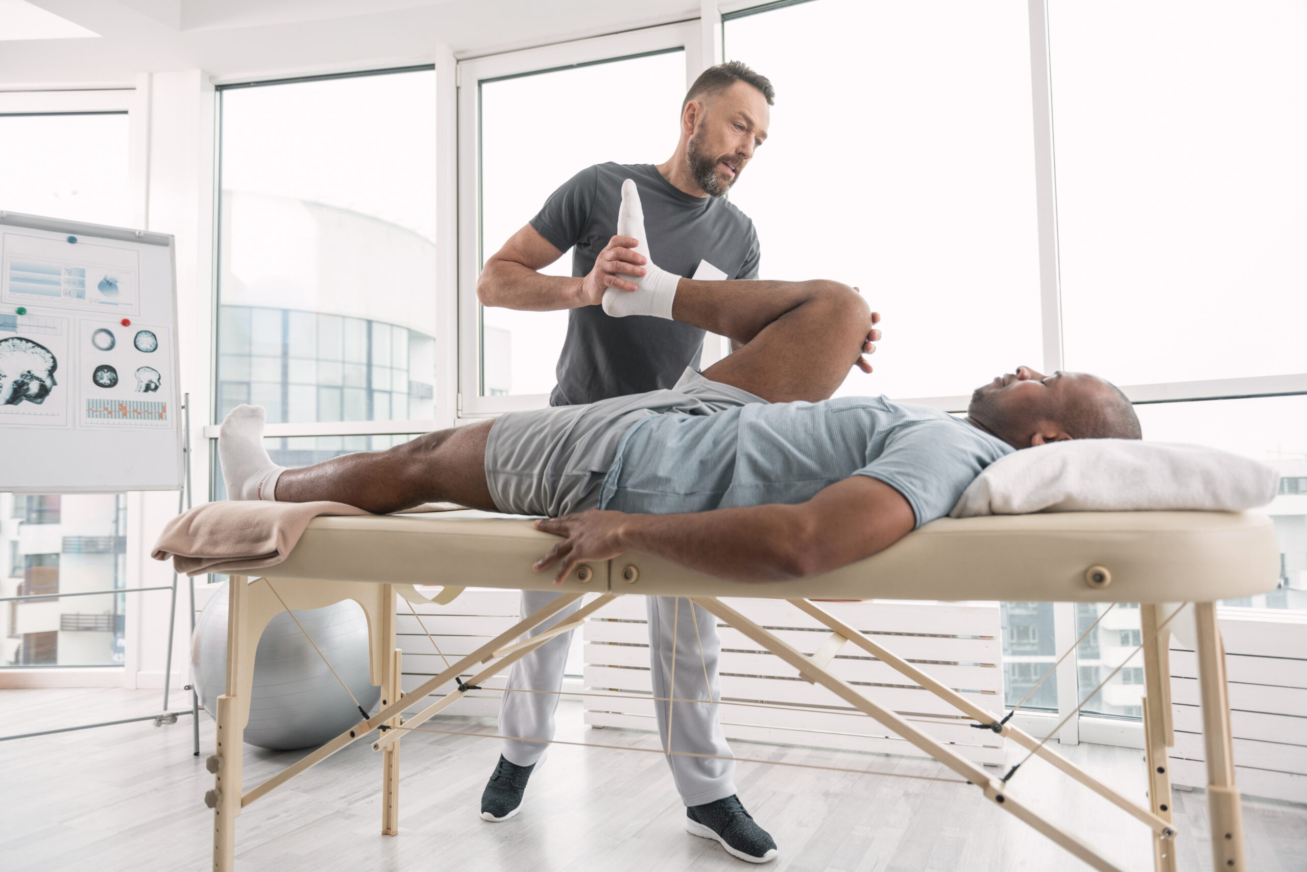 A male physical therapist bends a male sciatica patient's right leg to relieve back pain on a table in front of windows