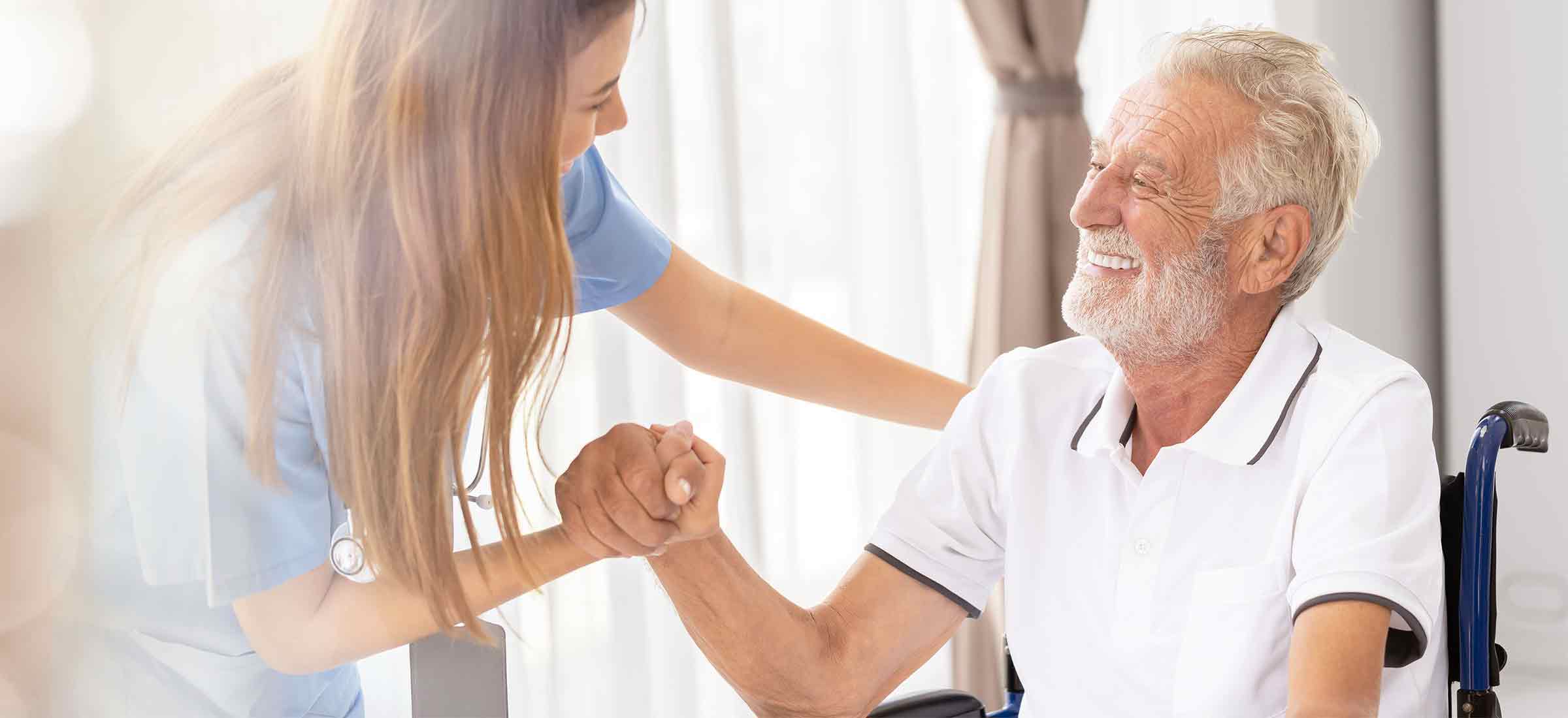 A nurse grabs a patient's hand as he sits in a wheelchair and smiles.