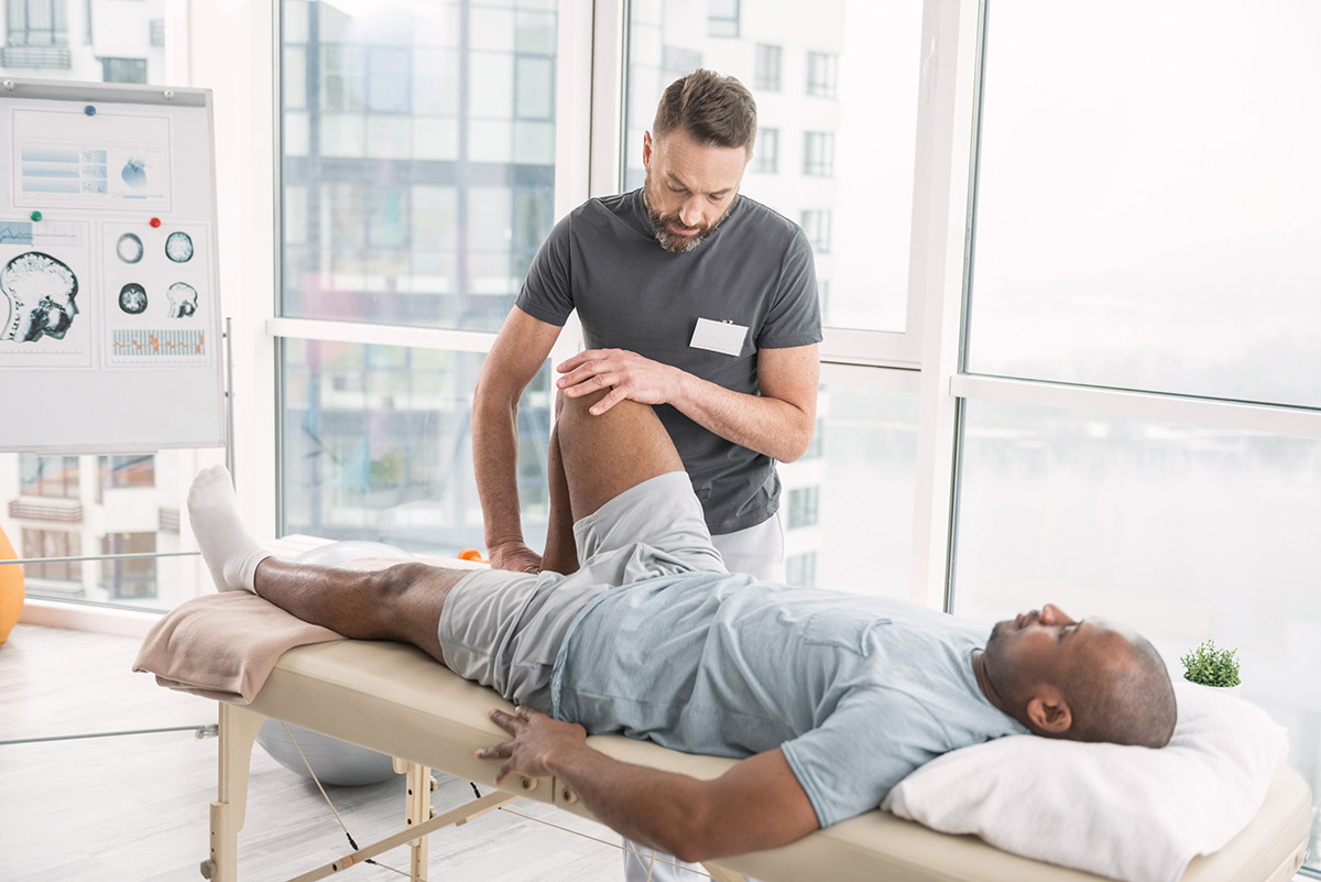 A male Keck Medicine physical therapist works on the bent knee of a man in shorts lying down in a medical setting by windows