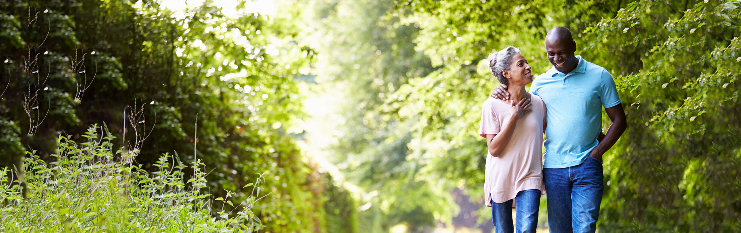 A senior woman with hearing aids walks with her arm around her husband outdoors among the trees