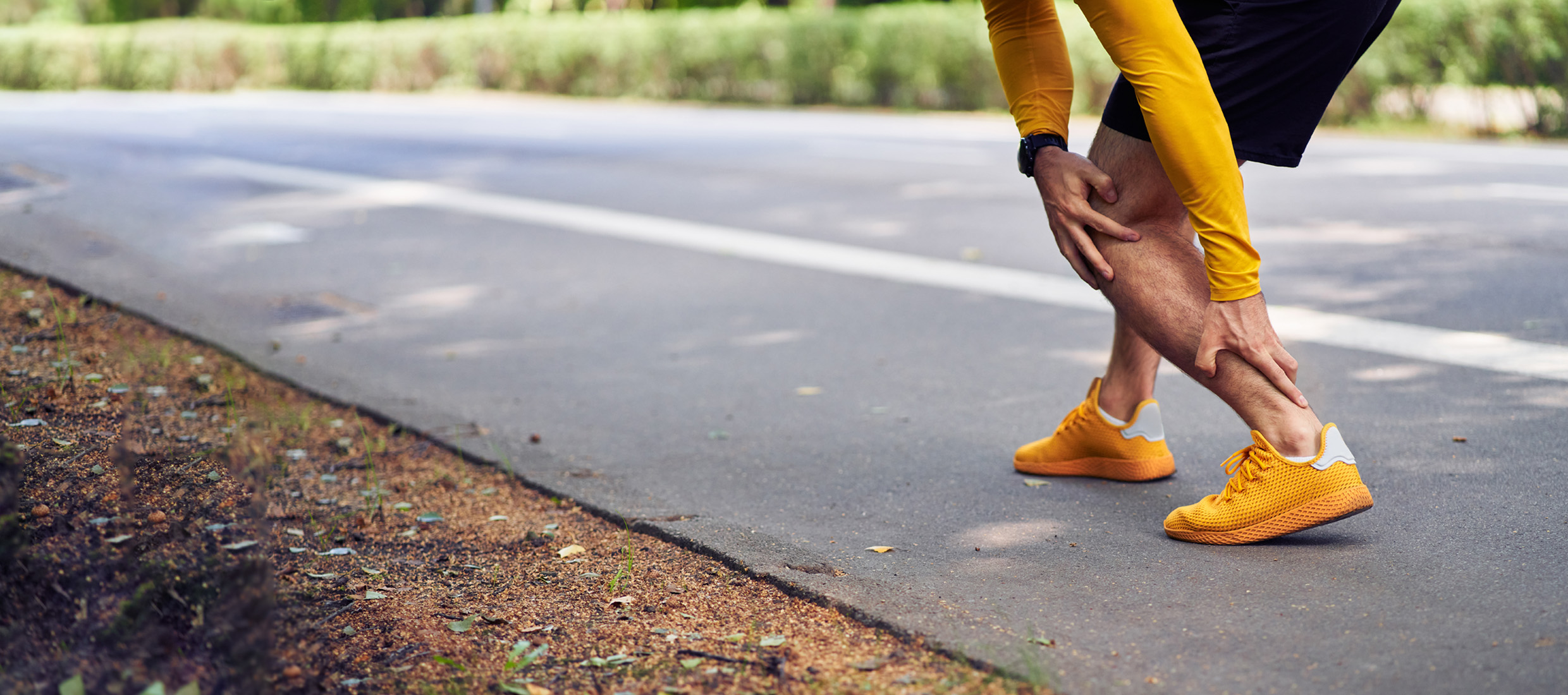 A man with a yellow shirt and shoes and black shorts has heel pain and holds his Achilles tendon on a country road