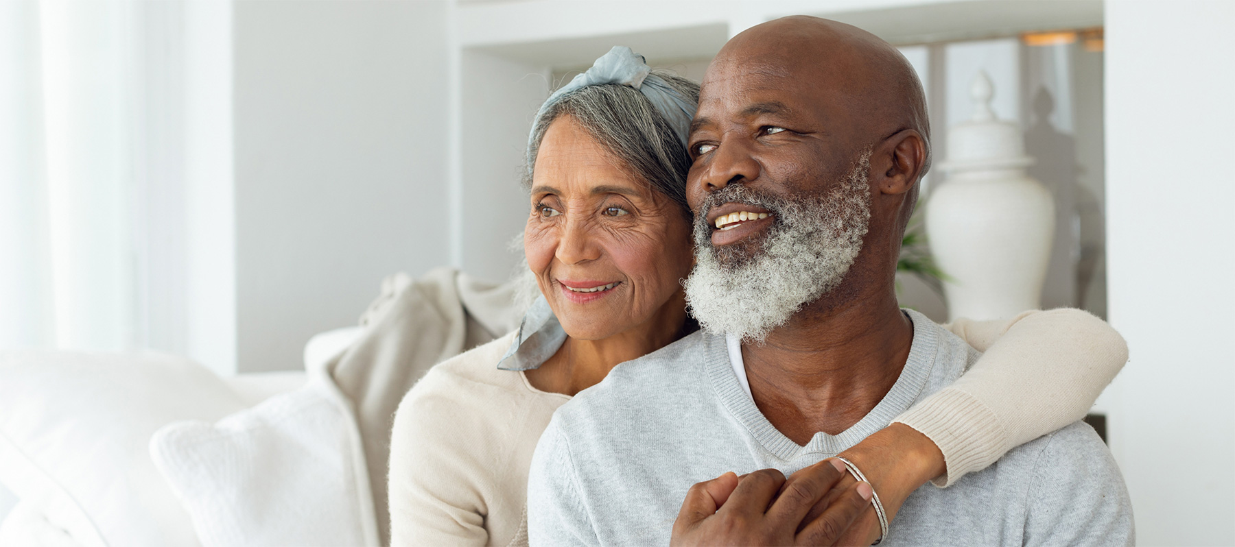 A senior woman stands behind a senior man with sleep apnea and hugs him with her left arm around his shoulder at home