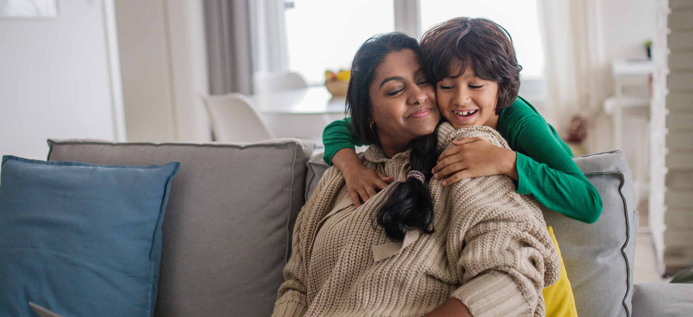 A kid hugs their mother's neck while she sits on a couch.