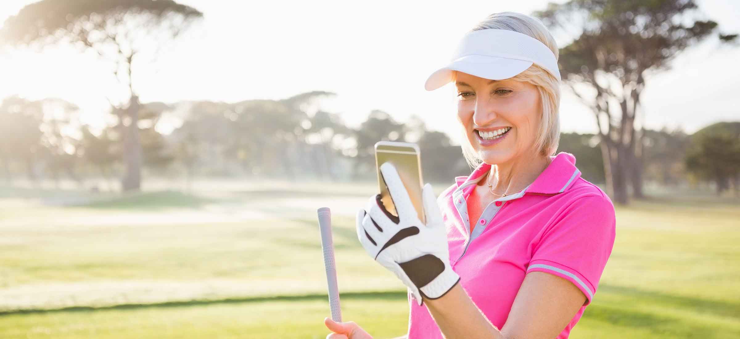 A woman smiles while looking at her phone on a golf course.