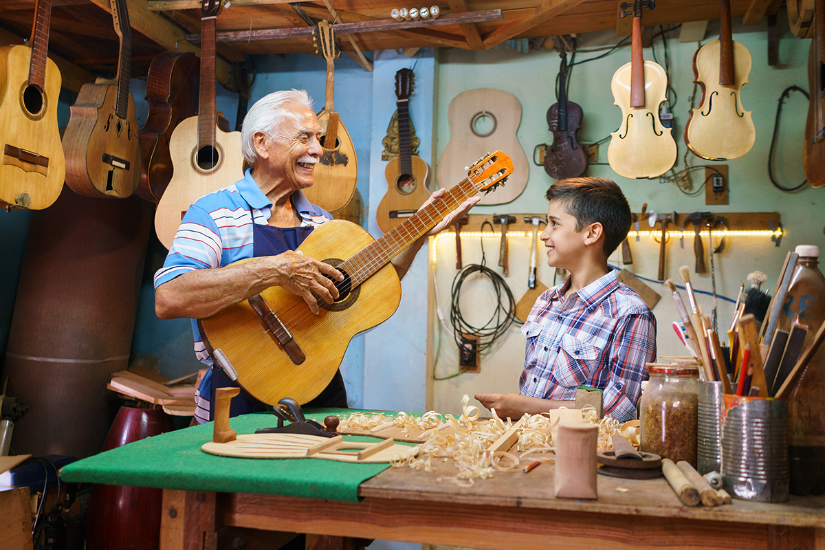 A senior male chordoma patient holds a guitar and smiles at a boy in his woodcarving shop with guitars on the walls