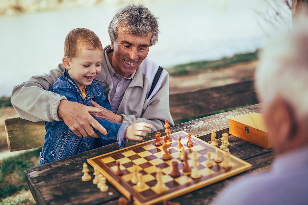 A male degenerative disc disease patient holds a smiling boy in a denim jacket who reaches for a chess piece on a chessboard