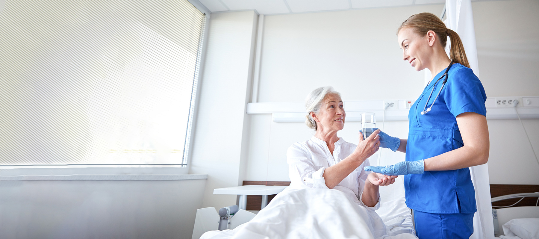A female Keck Medicine specialist with blue gloves hands a glass of water to a female senior patient with HIV in a white bed