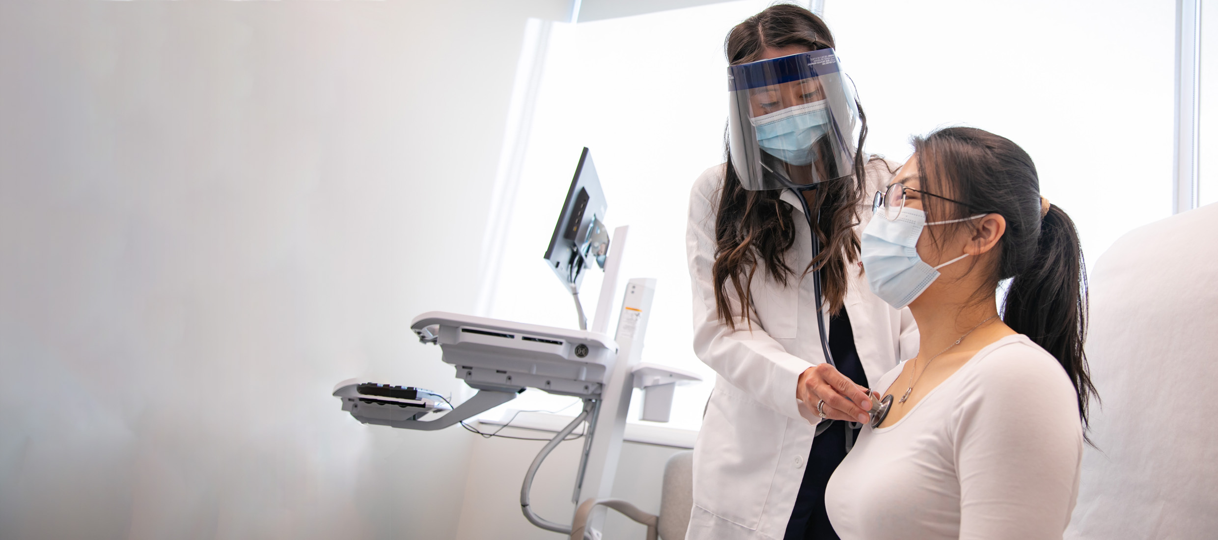 A female Keck Medicine oncology expert in a mask and face shield examines a female lung cancer patient with a stethoscope