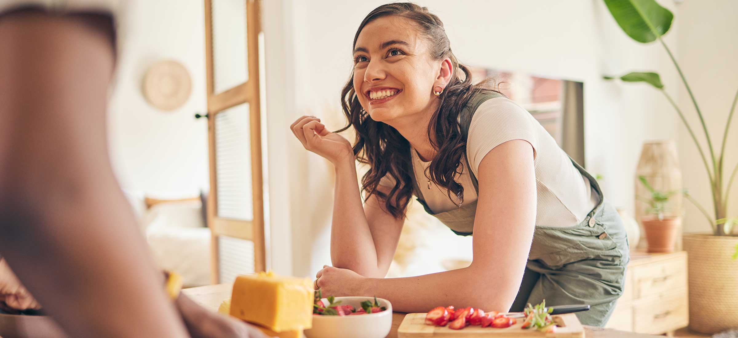 A woman bends over a kitchen table with food on it and smiles.