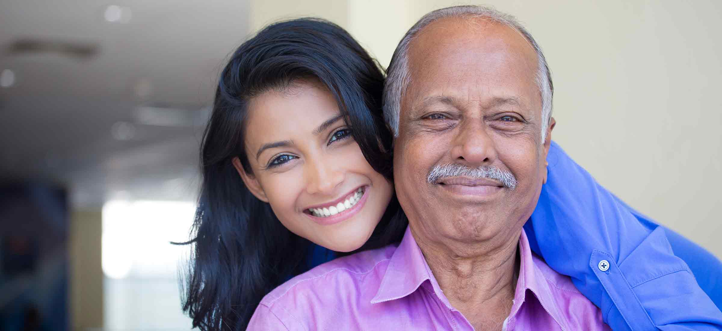 Closeup portrait of a young woman in blue shirt holding older man in a pink collared button down shirt, happy indoors with a home background.