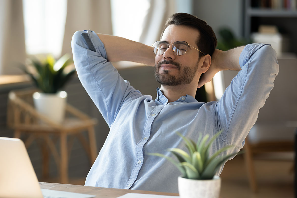 Calm millennial man in glasses sits relaxed at home office, distracted from computer work.