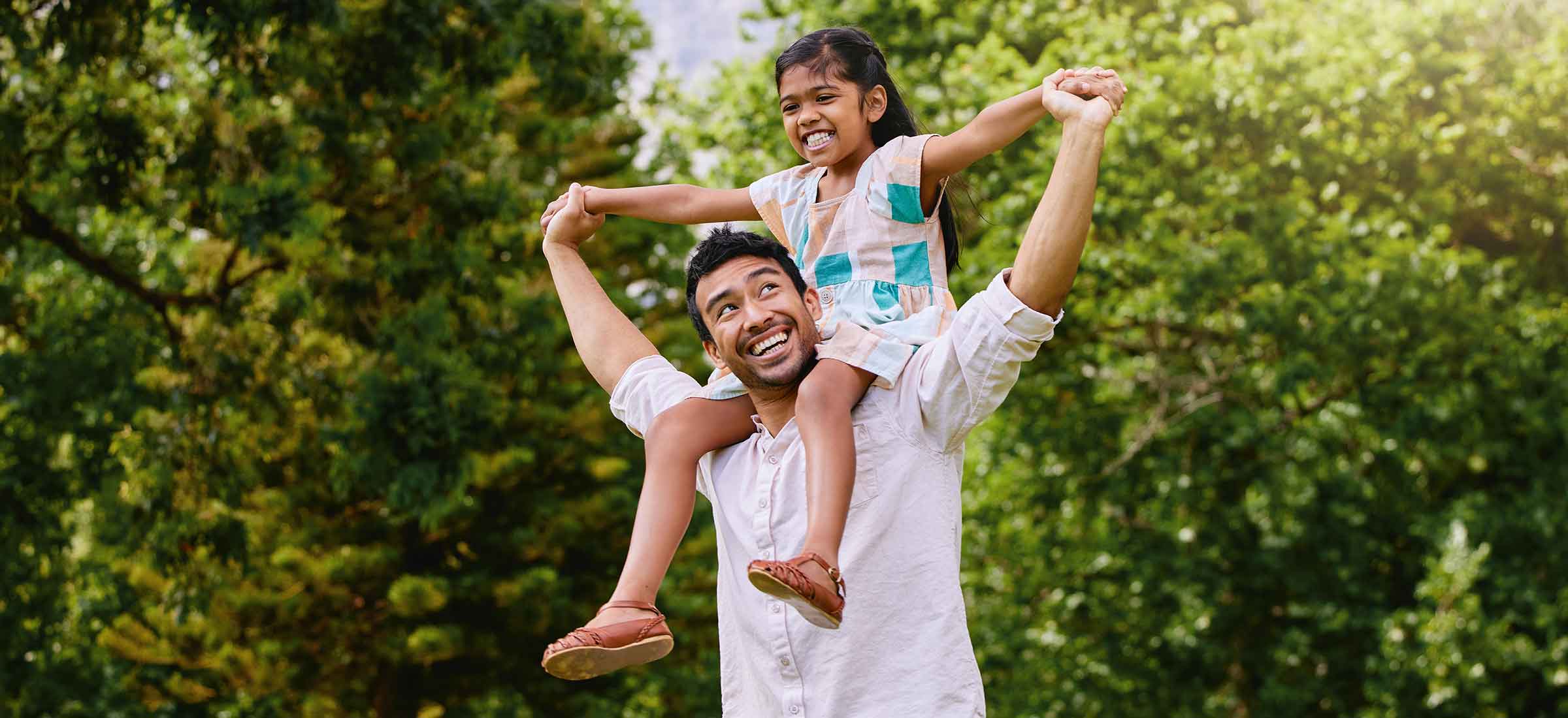 Healthy young man standing outdoors in park with his daughter on his shoulders.