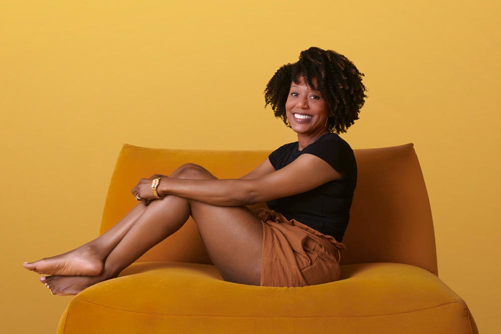 Candice Cauley, a Black woman in her thirties, wraps her arms around her knees on a burnt-orange couch and smiles for the camera