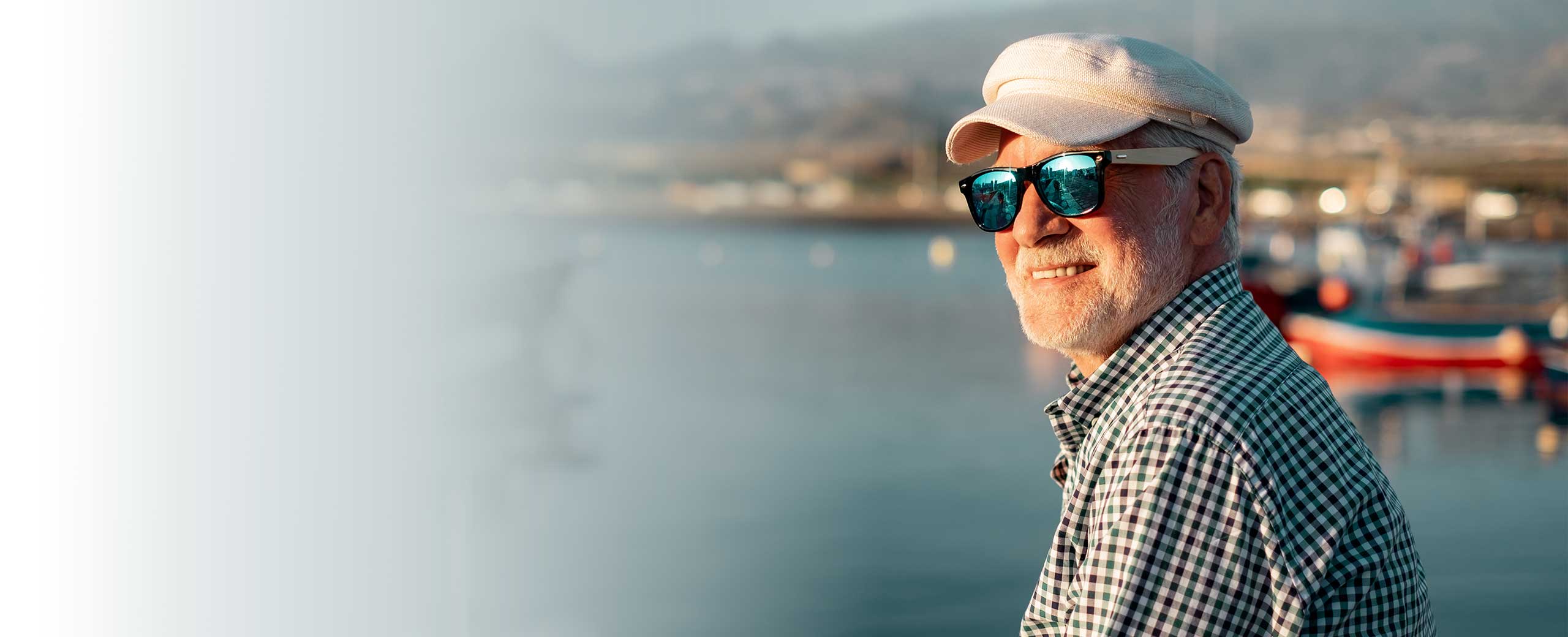 A man smiles on the coast while wearing sunglasses and a hat.