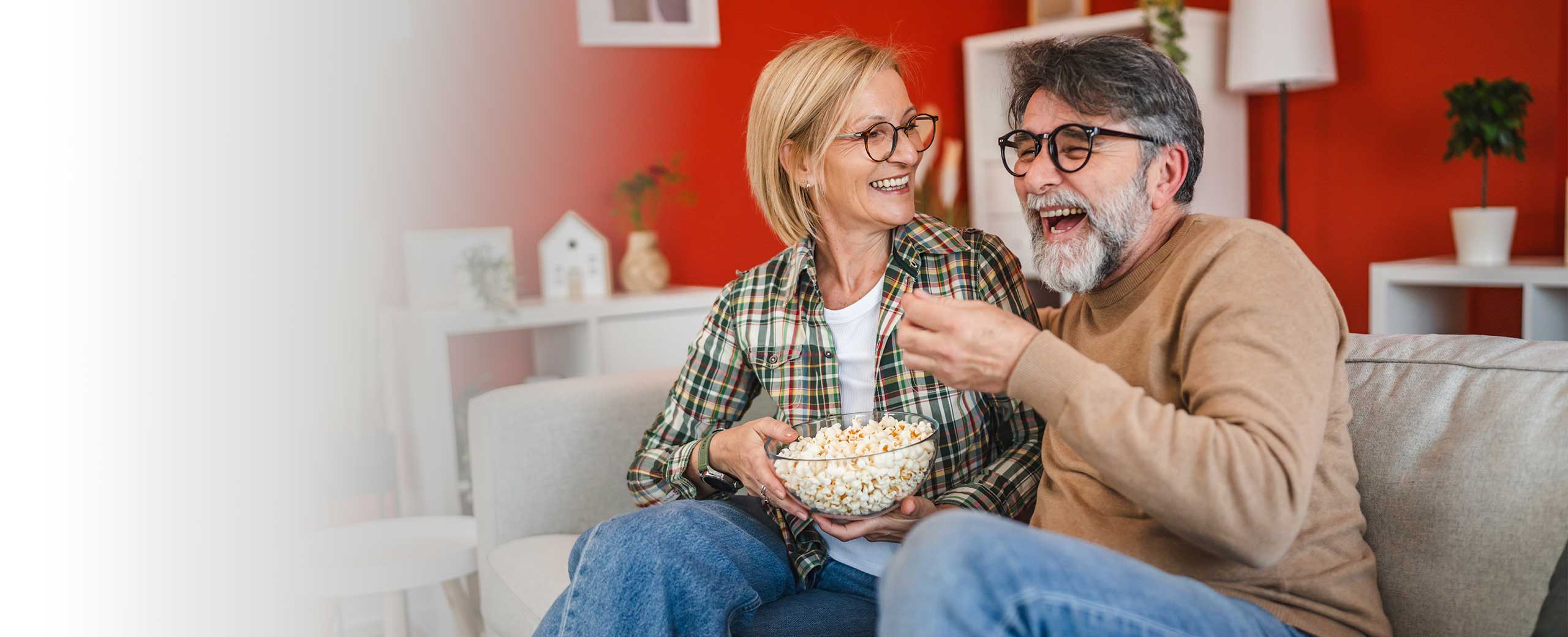 A man and a woman sit on a couch together holding a bowl of popcorn.