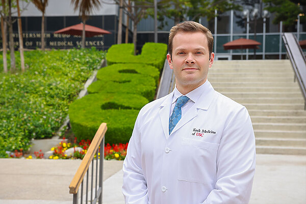 Dr. Collon stands in a white medical coat smiling in front of a set of stairs and shrubs.